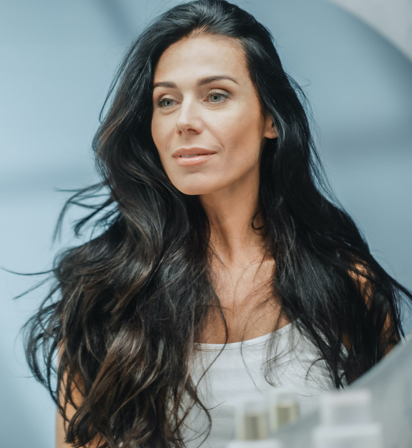 Photo of a middle-age woman with dark hair looking in the mirror at her skin